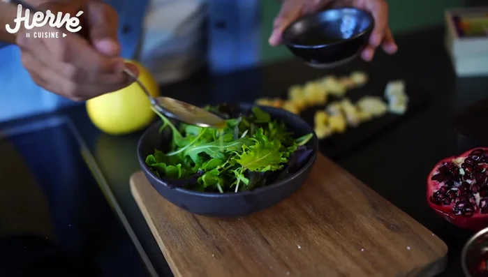 Assembler la salade : Mélanger les jeunes pousses, la roquette, les pommes, les graines de grenade, les cubes de Pont l'Évêque et Livarot, et la vinaigrette.