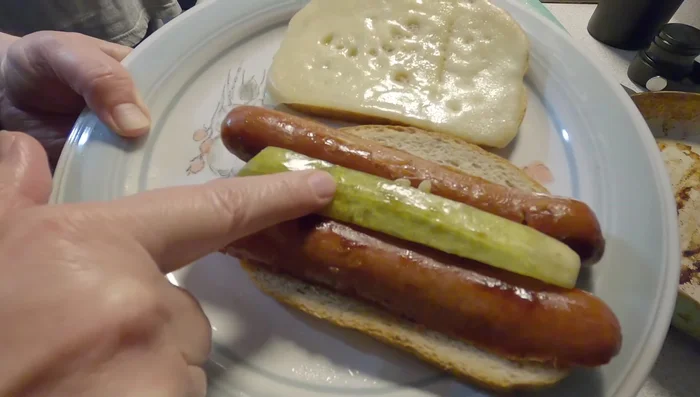 Préparation du sandwich (version mari) : Disposer les demi-saucisses et le cornichon sur le pain. Ajouter de la moutarde et du fromage suisse. Couper en deux.